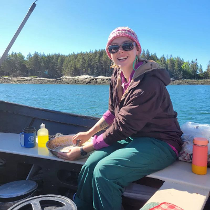 The image shows a woman on a boat, smiling and looking at the camera. She is wearing a hat, sunglasses, and a jacket. She is sitting on the edge of the boat, with a pan in her lap. The background shows a body of water and trees.